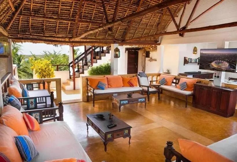 An open lounge under a makuti roof with carved daybeds, orange cushions and a view out to the palms
