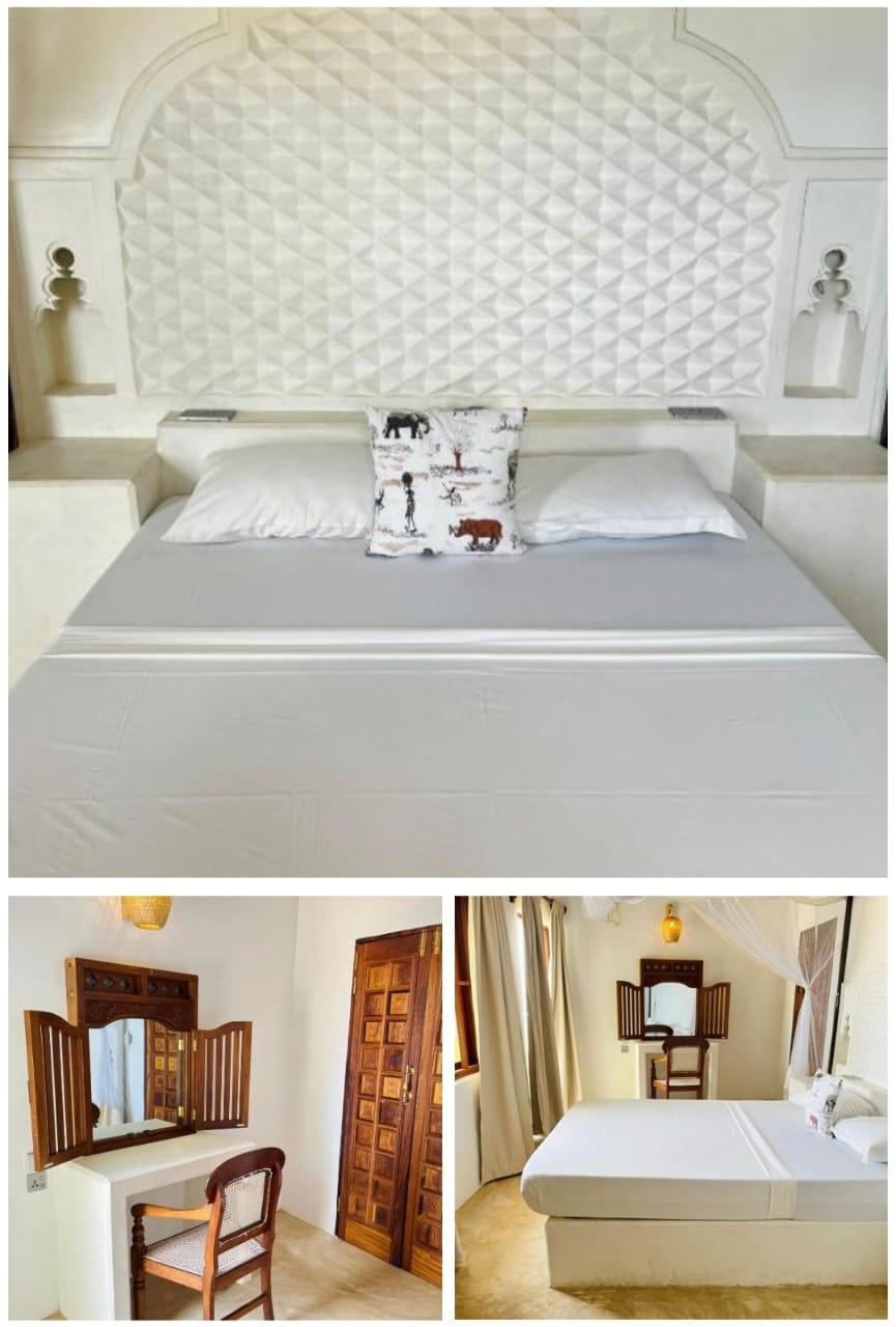 A wide bed against a moulded plaster headboard, a carved dressing mirror, and a second bedroom in white plaster