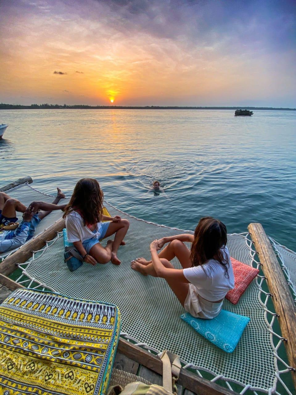 Guests sitting on a rope net slung over the water at sunset, one of them swimming below, the sun low over the creek