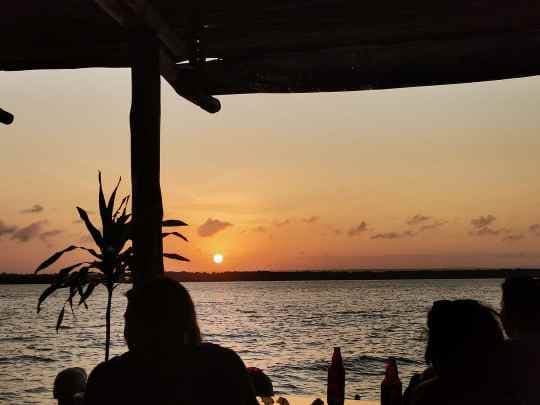 The sun setting over the water, seen in silhouette from under the palm-thatch roof of the stilt bar