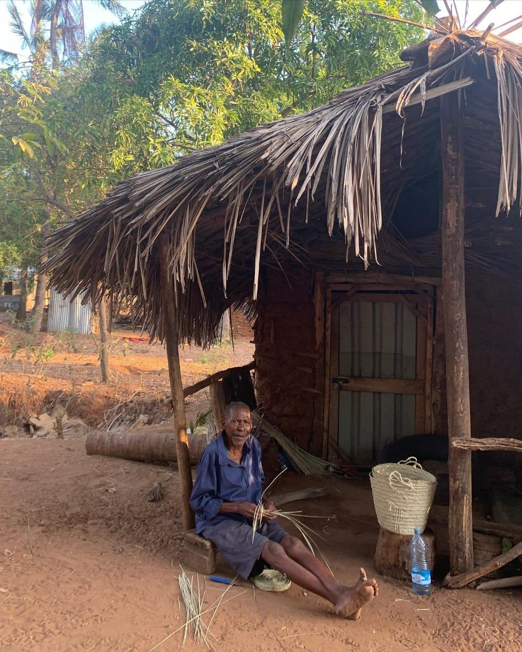 An elderly man sitting outside a mud-and-palm-thatch house, weaving palm leaf by hand on the red earth