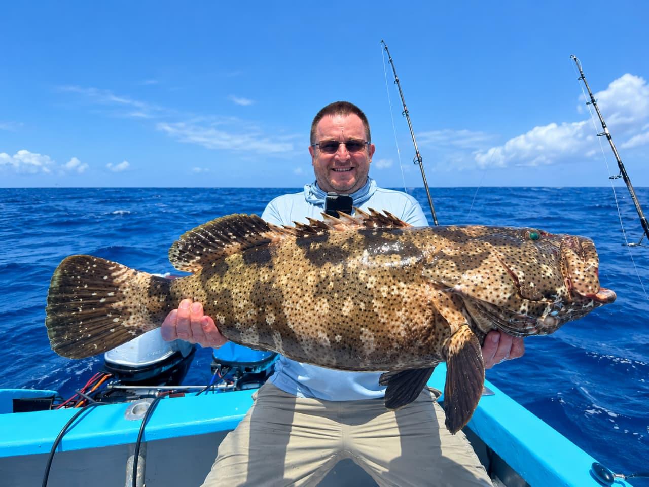 An angler on the bow holding a large spotted grouper in both arms, deep blue water behind