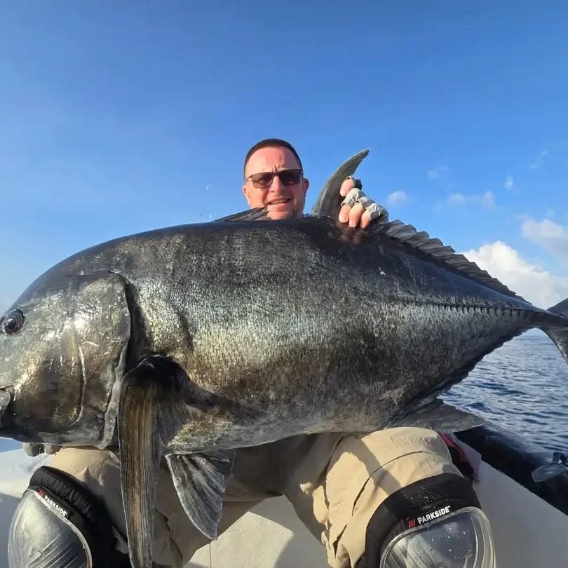 A dark, heavy-shouldered giant trevally held across an angler’s lap