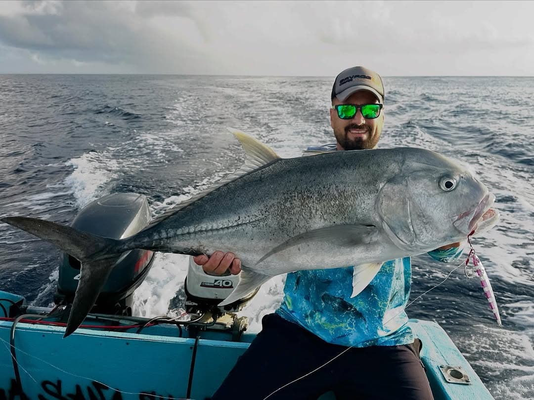 An angler at the stern holding a large giant trevally, the boat’s wake behind him