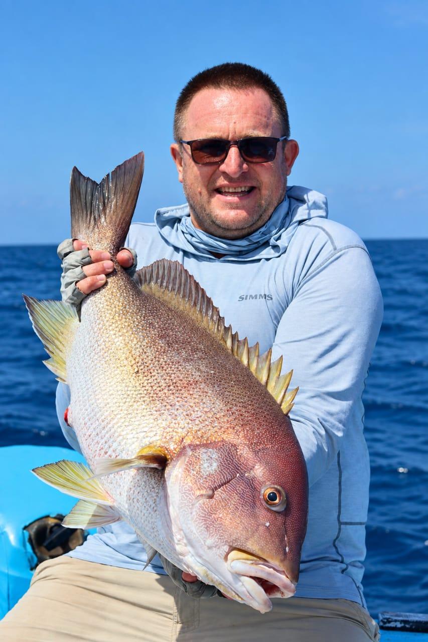 An angler holding a big rosy snapper upright against his chest, rods and open ocean behind
