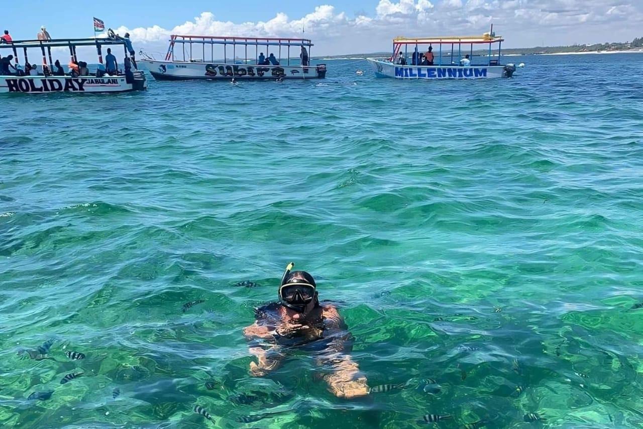 A snorkeller surfacing in shallow turquoise water with fish below and 3 boats moored behind