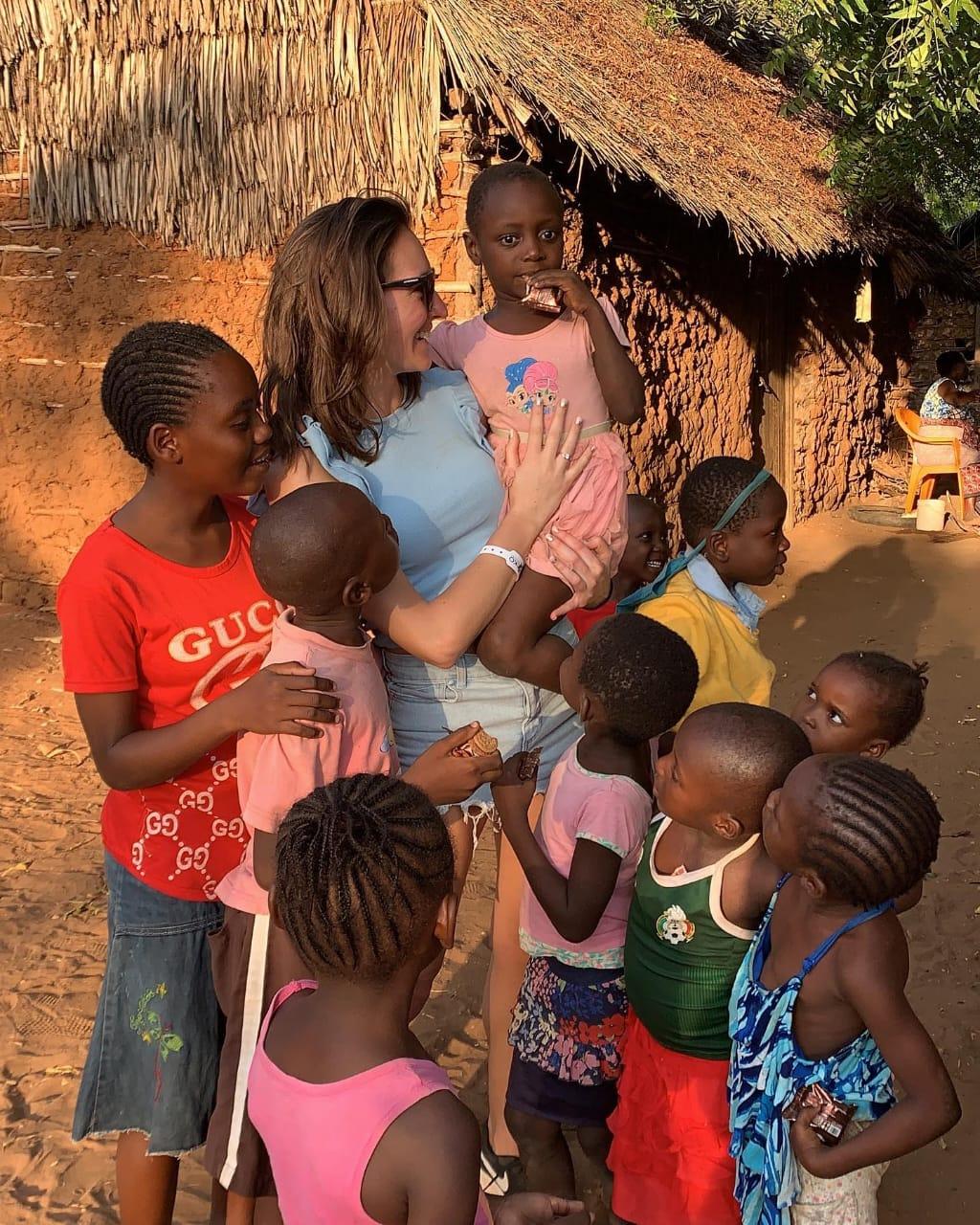 A visitor holding a small girl outside a village house, surrounded by children eating biscuits she has handed out